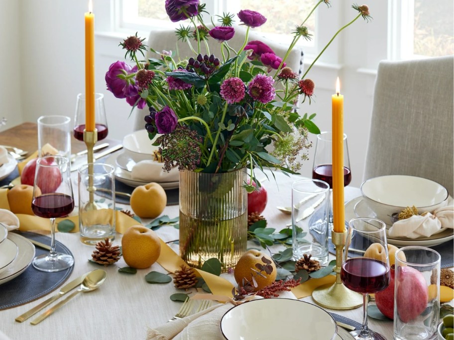 a dinging room table with place settings and fruit with a large glass ribbed vase / candle holder in the middle as a centerpiece with purple wildflowers in it