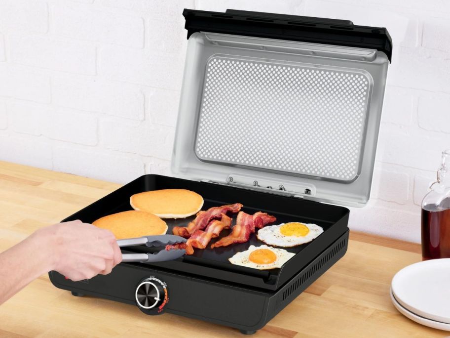 person using tongs to flip bacon on Ninja Sizzle 14" Electric Griddle