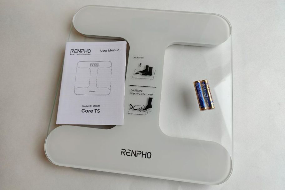 a glass bathroom scale shown with user manual and two AAA batteries