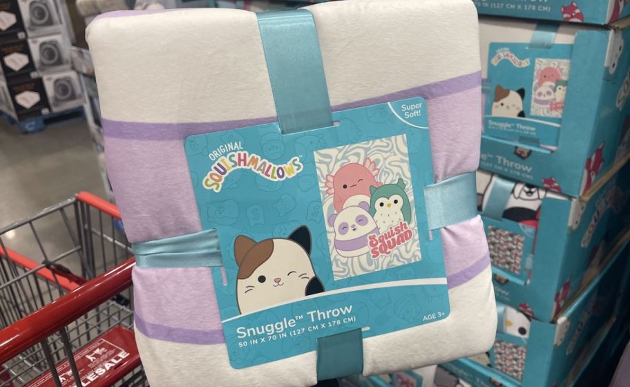 a womans hand holding a folded squishmallows throw blanket in a costco store