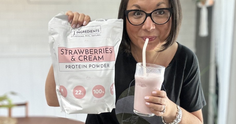 woman holding a bog of Strawberries & Cream protein powder while sipping on a straw from a glass with the protein powder