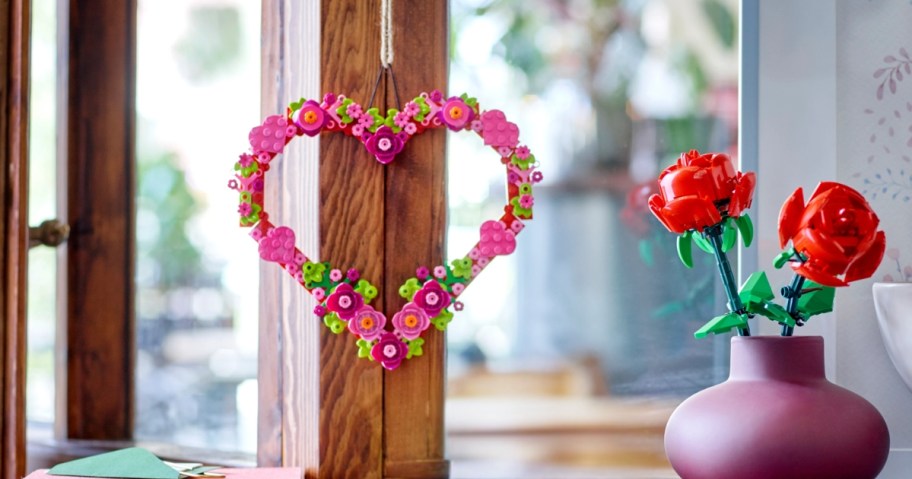 a LEGO pink, red and green heart shaped floral wreath ornament hanging on a window, purple vase with red LEGO roses next to it