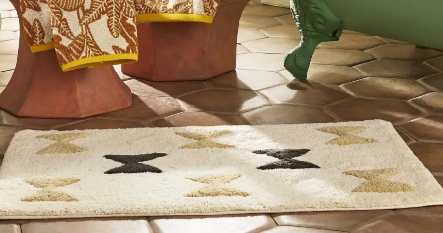 a cream bath rug with tan and black abstract designs on the floor in a bathroom