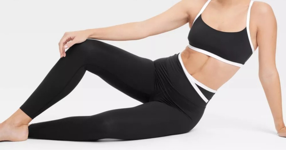 woman sitting her side sort of laying with 1 leg up - she's wearing activewear leggings and a sports bra in black with white piping details