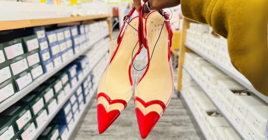 woman's hand holding a pair of red and clear high heel shoes showing the pointed toe with a heart shape design, a store aisle with more shoes behind them