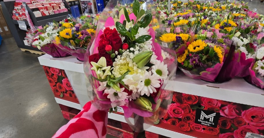 hand holding a bouquet of flowers with white lilies, white daisies, and red roses wrapped in plastic - a display with more flower bouquets is behind it