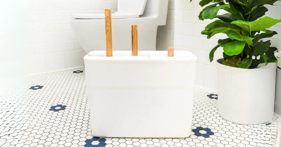 a white rectangle container with 3 handles sticking out, it's a bathroom trash can, toilet brush and plunger holder and set - sitting in a bathroom on white title floor, toilet in the background