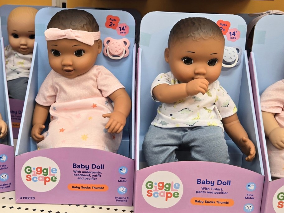 two baby dolls in boxes on a store shelf, one is a girl doll with brown skin and hair and one is a boy doll with brown skin and hair