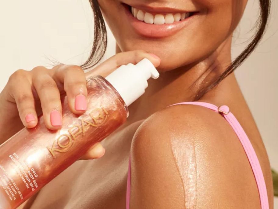 tan woman putting Kopari Beauty Rose Gold Sun Shield Body Glow SPF 45 5oz on her shoulder