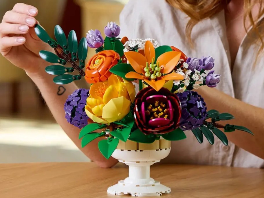  a woman sitting behind and reaching for a LEGO floral arrangement that's in a white LEGO vase