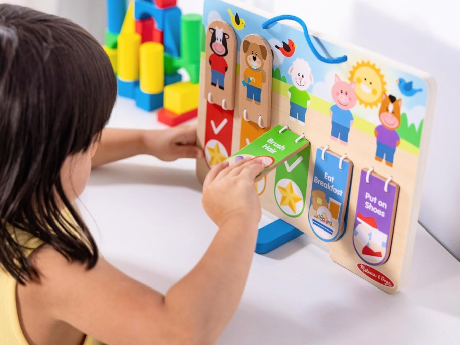 young girl with dark hair lifting the flap on a Melissa & Doug Wooden My Daily Routines Chart