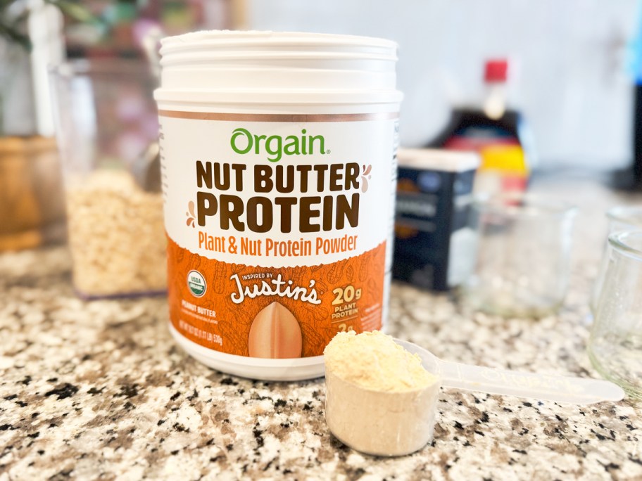 container of Orgain X Justin’s Organic Vegan Nut Butter Protein Powder with scoop on counter in front of it