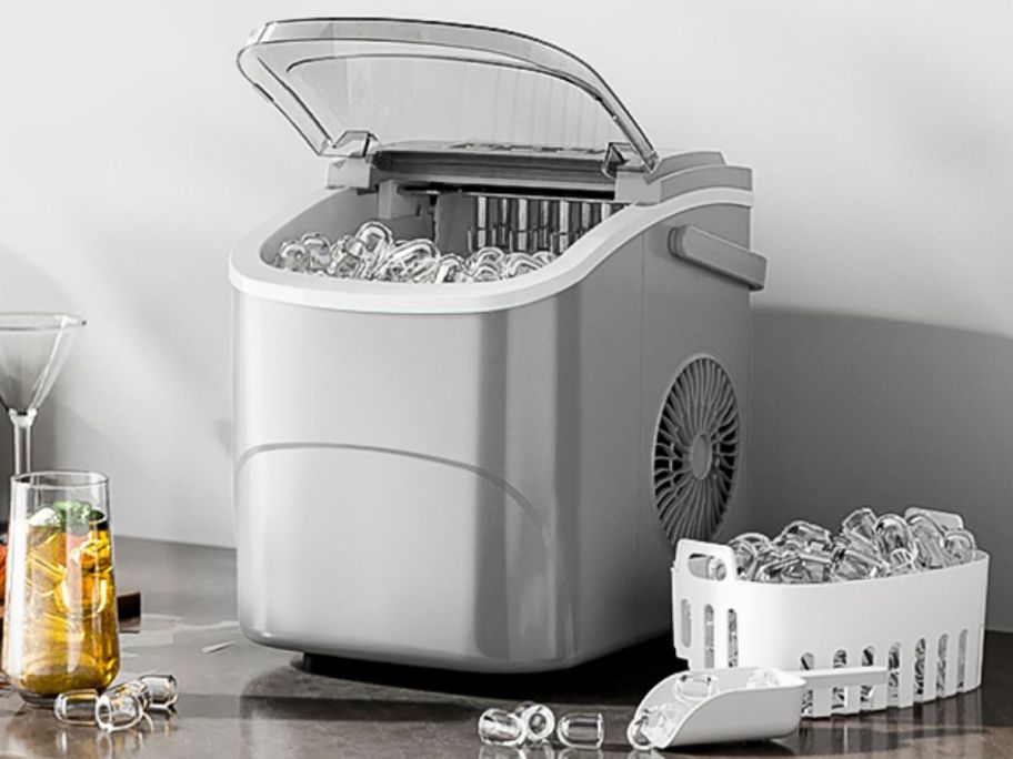 A silver ice maker on the counter surrounded by beverages and ice 