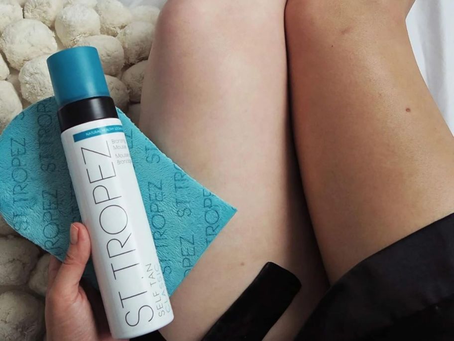 Woman holding a bottle of St.Tropez Self Tan Classic Bronzer Mousse Foam next to one white leg and one tan leg