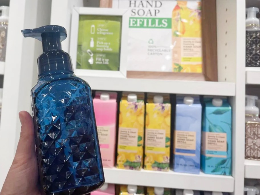 hand holding soap dispenser and refills behind it on shelf