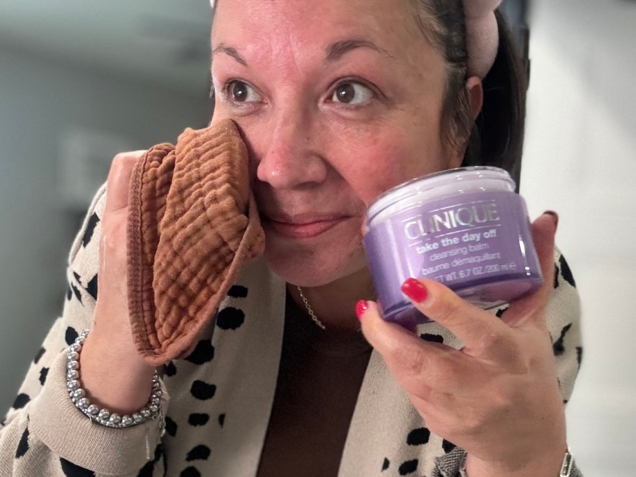 woman using a tan washcloth to wipe makeup off her face, she's holding a purple jar of Clinique makeup remover in her other hand