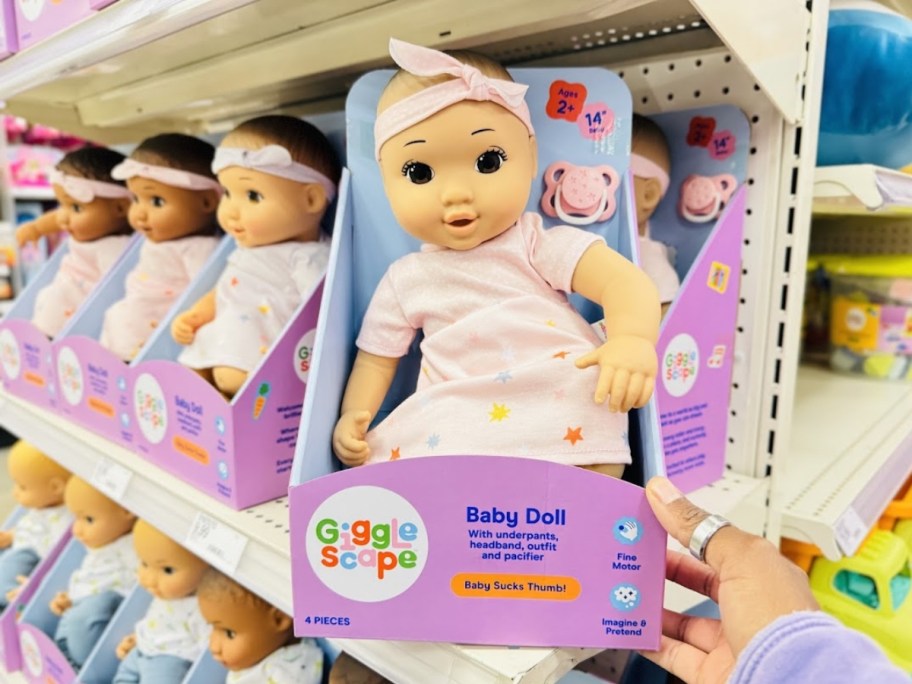 a baby doll with a white skin tone and brown eyes wearing a light pink dress with stars on it and a matching headband sitting in the packaging it onlinees in on a store shelf, more dolls behind it