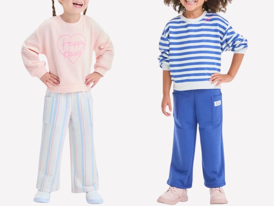 little girl wearing a peach sweatshirt with a pink heart that says "Happy Heart" and matching striped pants, and a little girl wearing a blue and white striped top with red hearts on the shoulder and matching blue pants