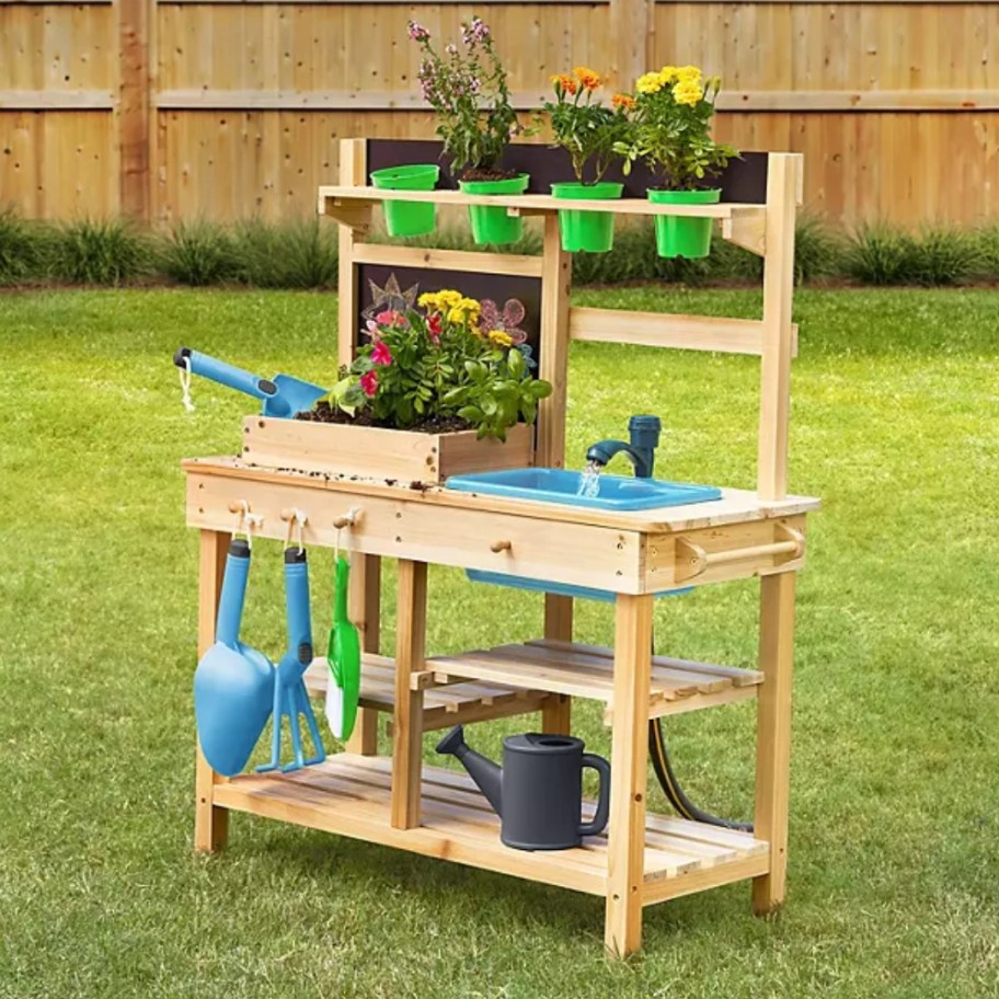 a kid's wooden outdoor play kitchen with plants on top and gardening supplies on it