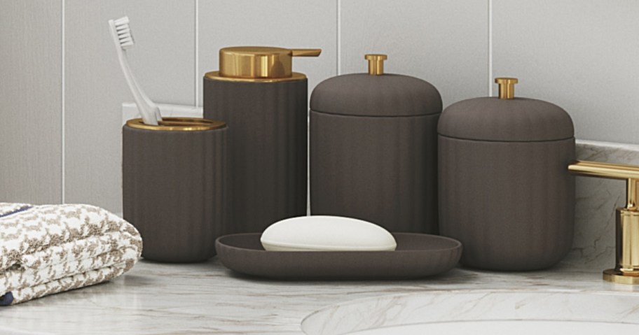 a set of bahtroom accessories on a bathroom counter next to a sink. The items are a dark grey with a ribbed design and gold accents and includes a toothbrush holder, soap pump, vanity tray, and 2 jars with lids.