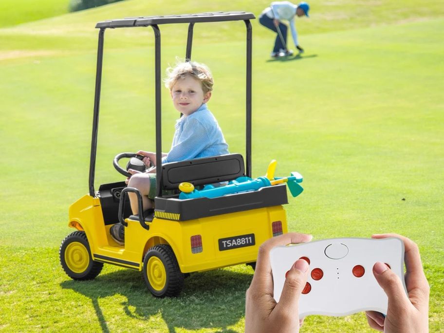 Kids riding in an electric golf cart on grass with an adult's hands using a remote control nearby