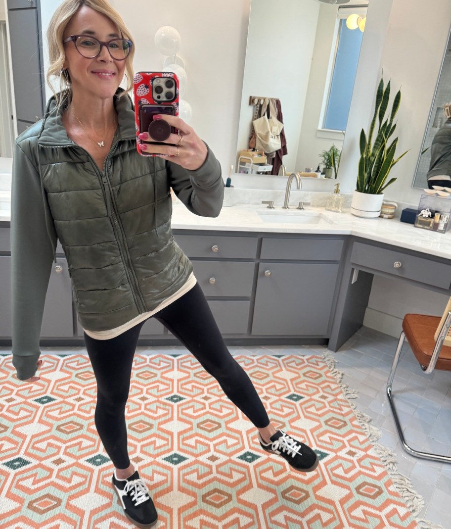 woman taking selfie in mirror wearing black leggings and green puffer coat 