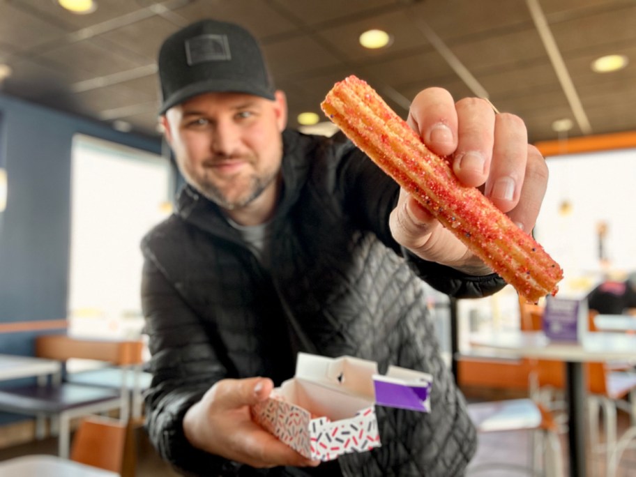 smiling man wearing a hat holding a milk bar birthday cake churro inside a taco bell restaurant