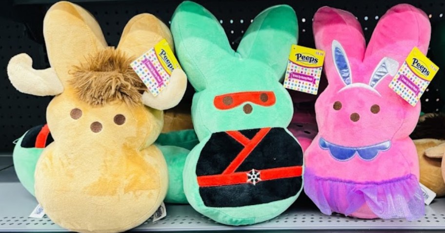 3 plush Peeps bunnies on a Walmart store shelf, one is green and dressed in black and red Ninja gear, one is pink and dressed in purple ballerina outfit, and one is tan and has horns and a tuft of hair so he looks like a Buffalo