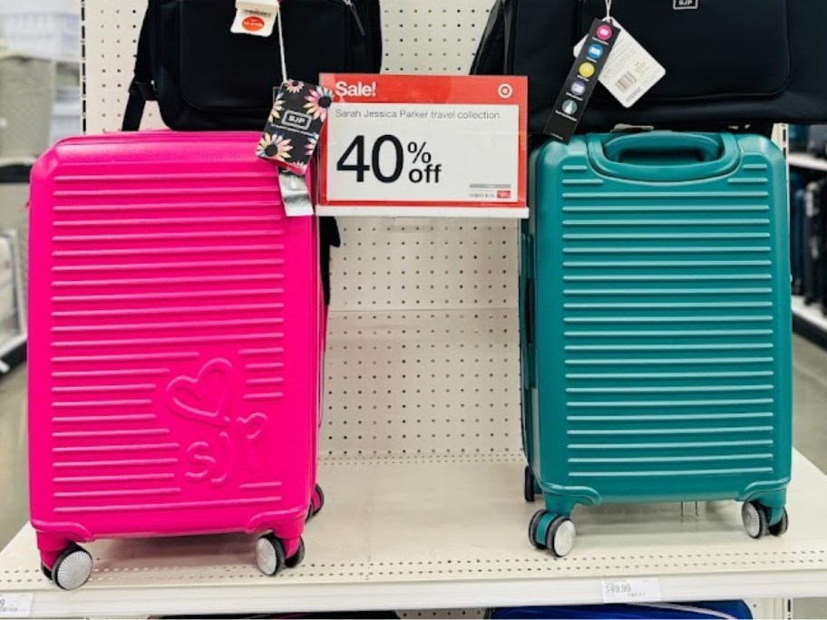 2 hardside spinner carryon size suitcases next to each other on a store display endcap with a sign that says 40% off - one is a a teal/emerald green color and the other is a hot pink color