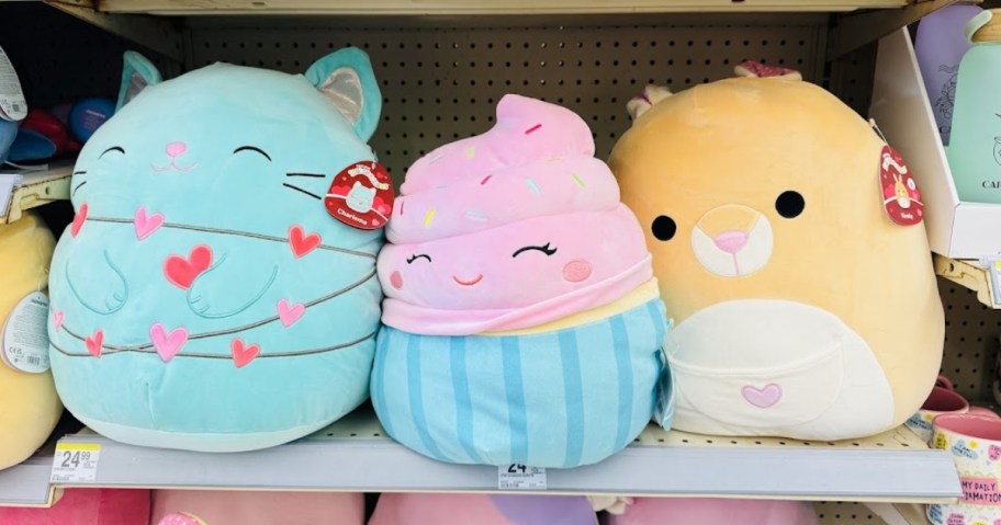 3 Valentine's Themed Squishmallows on a store shelf, one is a green cat with a string of hearts, one is a cupcake with pink icing and one is a dog