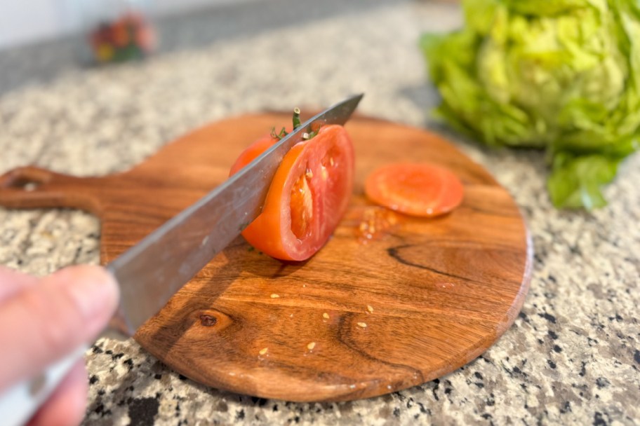 knife cutting tomato on wood cutting board next to lettuce