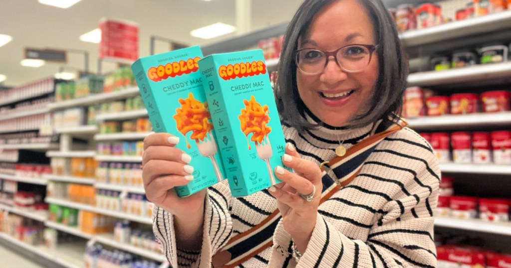 Woman in store aisle, holding up two boxes of Goodles protein, Mac and cheese