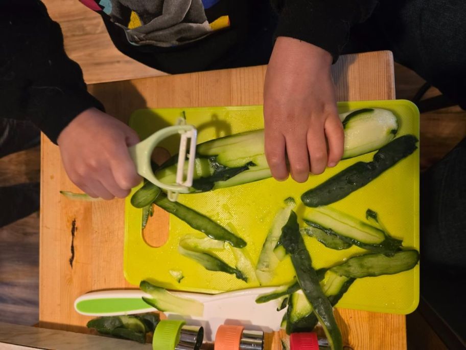 child using peeler to peel cucumber on cutting board