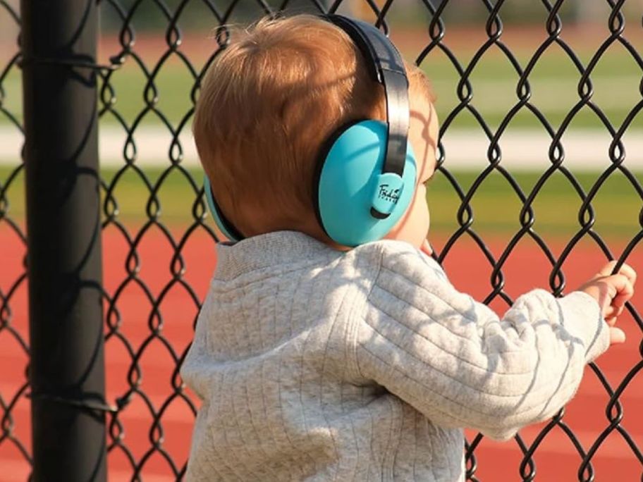 Little boy watching a sports game from behind a chain-link fence wearing noise-canceling headphones