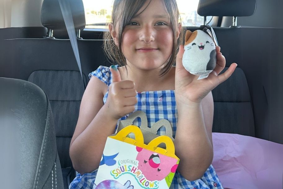 a little girl holding a squishmallows happy meal toy and happy meal box