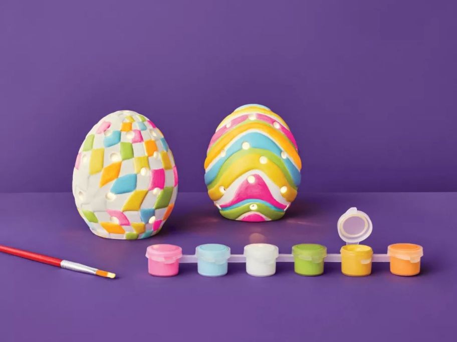 Mondo Llama Easter Ceramic Egg Painting Kit 2-Count painted with paints and brush in front of them against purple background