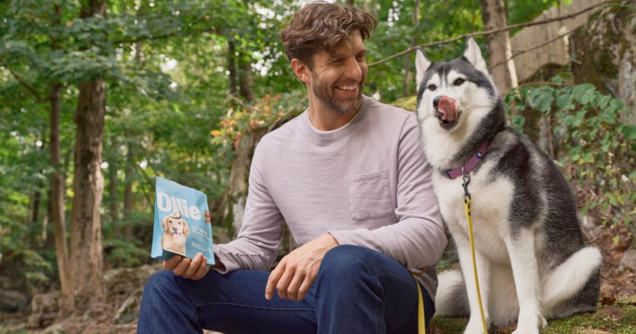 man holding a bag of ollie jerky strips sitting next to a dog