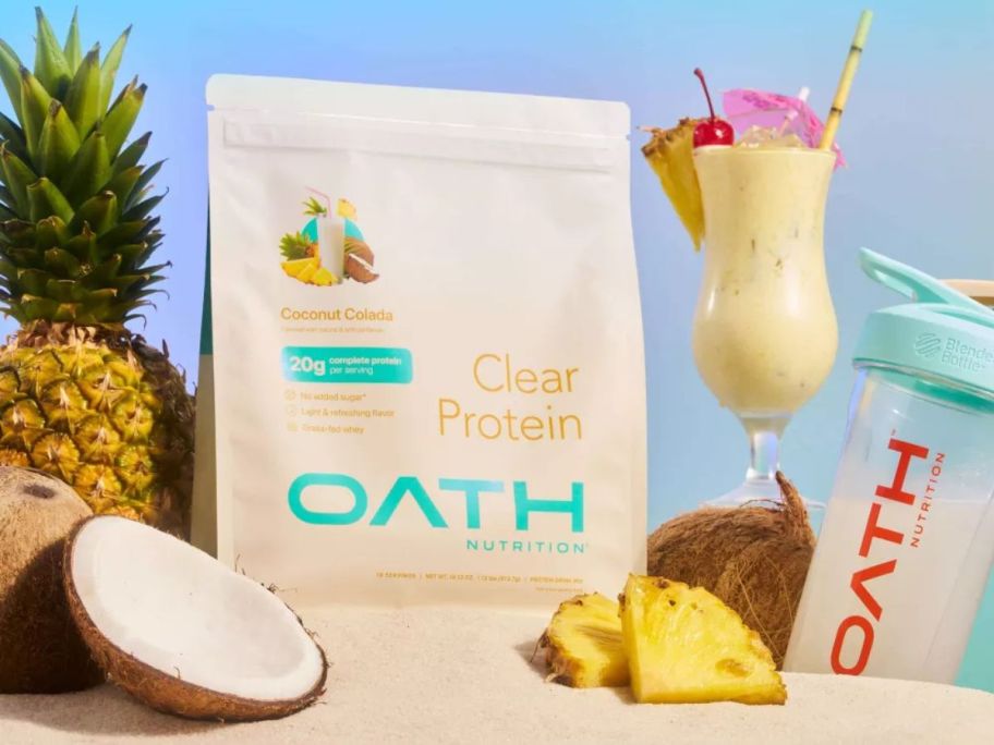 oath protein powder bag on sand amongst pineapples, coconuts, and a drink glass