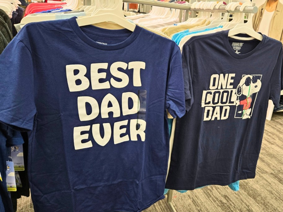 2 Dad t-shirts hanging on a store rack, one is blue and says "Best Dad Ever", the other is blue and has Snoopy and says "One Cool Dad"