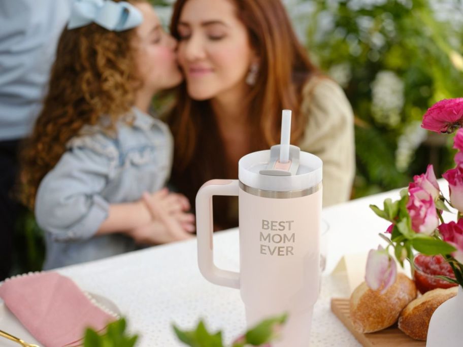 A Pink Stanley Tumbler on a table with Best Mom Ever Engraved on it with a little girl kissing a woman on the cheek in the background