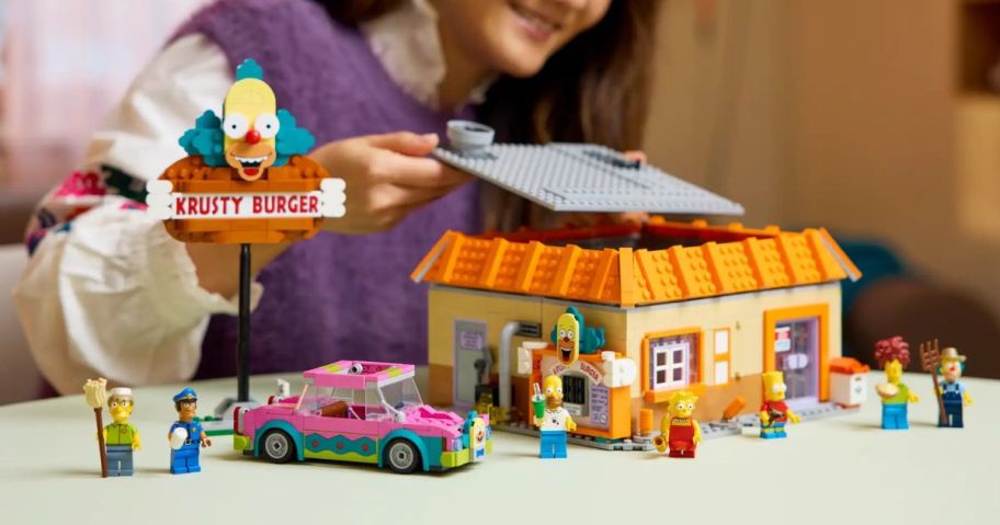 A woman playing with a Simpsons Krusty Burger LEGO set on a coffee table