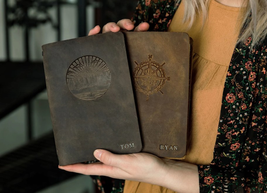 woman holding two leather journals with outdoor engravings on front