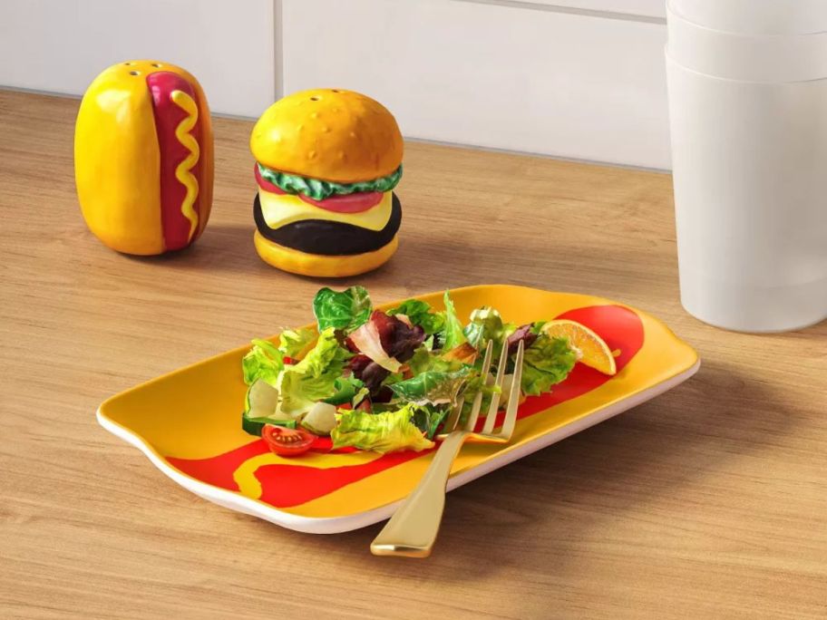 Room Essentials Hot Dog Serving Platter with salad and a fork on it with a hot dog and hamburger salt and pepper shaker set behind it on table